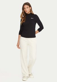 Top nero a maniche lunghe con collo alto e logo bianco. Abbinato a pantaloni bianchi a gamba larga e sneakers di colore chiaro. Tessuto liscio.