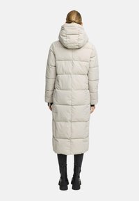 Persona con capelli biondi raccolti in una coda di cavallo, indossa un lungo cappotto imbottito beige chiaro con cappuccio, pantaloni neri e stivali neri alla caviglia, di spalle.