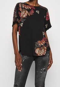 Svart kortärmad topp med blommönster i rött, gult och blått. Har en avslappnad passform och delikat broderi längs axlarna.
