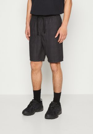 Homme portant un short à carreaux noir, des chaussettes noires et des chaussures de sport noires, se tenant devant un fond blanc uni.