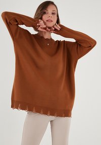 Maglione marrone oversize con scollatura a V, polsini a coste e orlo a onda. Texture liscia, realizzato in materiale lavorato a maglia.