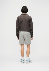 Άνδρας με σκούρα κυματιστά μαλλιά στέκεται πλάτη, φορώντας ένα καφέ καρό πουκάμισο με μακριά μανίκια, ανοιχτό γκρι σορτς, λευκές κάλτσες και μπλε-λευκά αθλητικά παπούτσια.