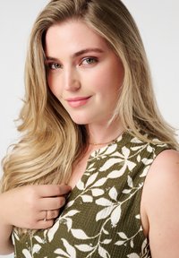 Groene textuurstof met wit bladpatroon; model draagt een delicate gouden ketting en ring, met zachte, golvende blonde haren.