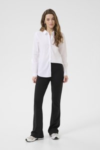 Chemise blanche à boutons avec un col classique, associée à un pantalon noir évasé et des baskets grises, sur un mannequin devant un fond uni.