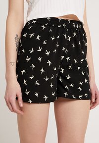 Shorts noirs en tissu léger avec une taille élastique, ornés d'un motif de oiseaux blancs sur l'ensemble. Texture lisse.