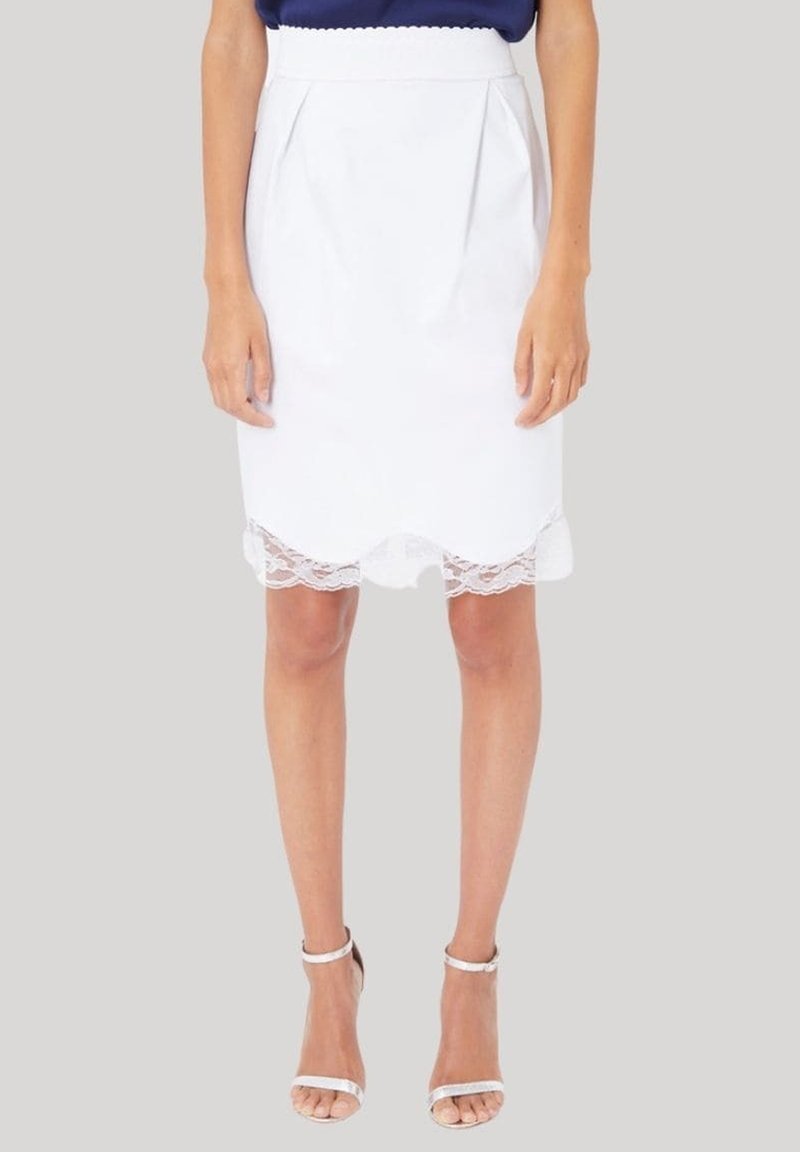 Mujer con una falda blanca hasta la rodilla con un dobladillo de encaje festoneado y sandalias de tacón alto con correa en el tobillo, contra un fondo sencillo.