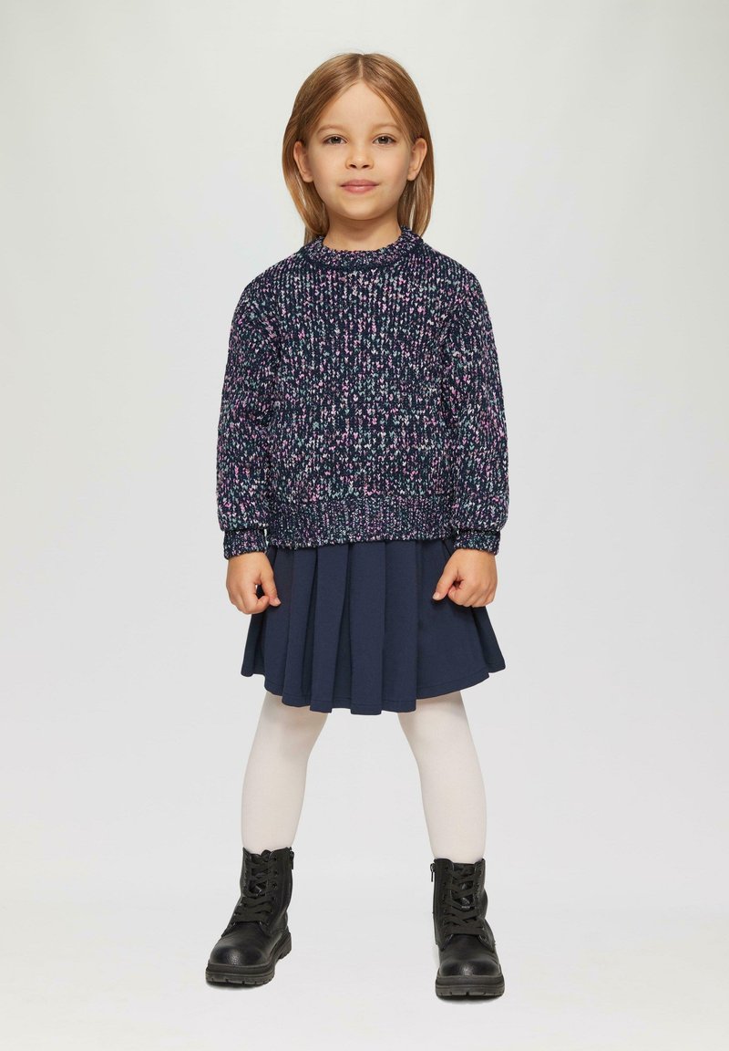 Dunkelblauer Strickpullover mit rosa Akzenten, plissierter marineblauer Rock, weiße Strumpfhose und schwarze ankle boots. Einfaches, geschichtetes Outfit für Kinder.