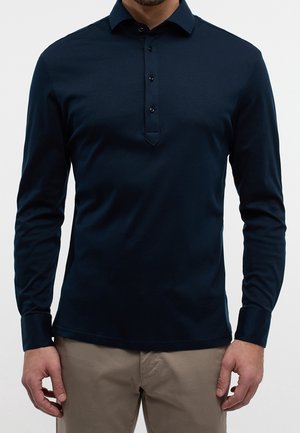 Uomo che indossa una polo a maniche lunghe blu navy con abbottonatura e pantaloni beige, in piedi davanti a uno sfondo uniforme.