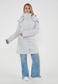 Donna con cappotto invernale grigio chiaro con cappuccio, jeans blu e sneakers bianche, in piedi davanti a uno sfondo bianco uniforme.