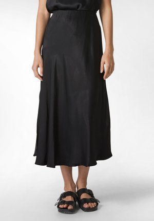 Schwarzer Satin-Midi-Rock mit fließender Silhouette, elastischem Taillenbund und seitlichen Schlitzen. Kombiniert mit schwarzen Sandalen mit offenen Zehen und verstellbaren Riemen.