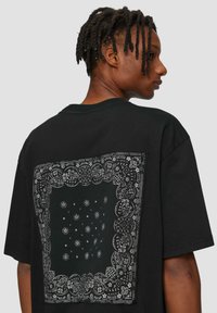Zwart T-shirt met een grote witte bandana-print op de achterkant, versierd met ingewikkelde bloemenpatronen en ronde accenten. Casual pasvorm.