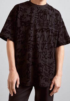 Mann trägt ein locker sitzendes schwarzes T-Shirt mit verschiedenen schwarzen Graffiti-Wörtern und Symbolen, die überall darauf gedruckt sind.