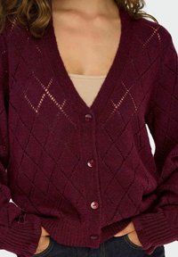 Cardigan en tricot bordeaux avec un motif en losange, décolleté en V et cinq boutons. Les manches sont côtelées aux poignets, ajoutant de la texture.