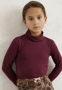 Bordo megztinis su aukštu kaklu ir ilgomis rankovėmis, derinamas su raštuotomis rudomis kelnėmis ir šviesiai rudoku diržu su metaline sagtimi.