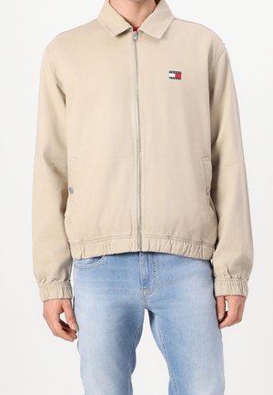 Veste beige zippée avec poignets et ourlet élastiques, petit logo Tommy Jeans sur la poitrine gauche, portée avec un jean bleu clair sur un mannequin masculin.