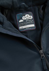 Veste imperméable marine avec un intérieur doux en polaire, arborant une étiquette colorée avec des nuages et des gouttes de pluie. Fermeture éclair à l'avant.