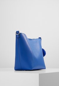 Borsa a mano in pelle blu con una consistenza morbida, caratterizzata da una silhouette arrotondata e un accento di pompon blu morbido sul lato.