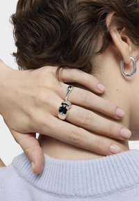 La bijouterie en argent présente un design en émail noir en forme de trèfle sur une bague, avec une forme courbée. Une boucle d'oreille en argent ouverte est portée, mettant en avant une finition épurée.