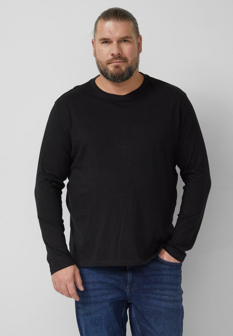 Schwarzes langärmeliges T-Shirt aus Baumwolle. Rundhalsausschnitt, lockere Passform. Kombiniert mit blauen Jeans, steht vor einem einfarbigen Hintergrund.