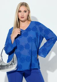 Blå bluse med lange ærmer og blomsterbroderi, V-udskæring, kombineret med en metallic sølvhåndtaske og blå bukser. Blødt, tekstureret stof.