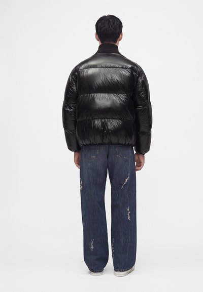 Personne aux cheveux courts et foncés porte une doudoune noire brillante et un jean bleu délavé et ample, debout de dos sur un fond blanc uni.