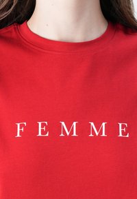 Rød bomulls T-skjorte med rund hals og hvit tekst som leser "FEMME" i fet, stor serif-skrift. Glatt tekstur.