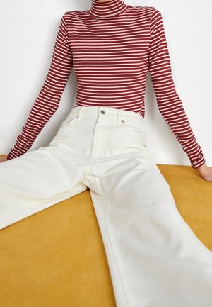 Person trägt roten und weißen gestreiften Rollkragenpullover mit langen Ärmeln und weißer Hose mit hohem Bund, sitzt auf einer senfgelben Oberfläche.