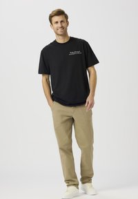 Fekete pamut póló, mellkasán nyomtatott szöveggel, khaki nadrággal és fehér tornacipővel párosítva, álló férfi alakon viselve.