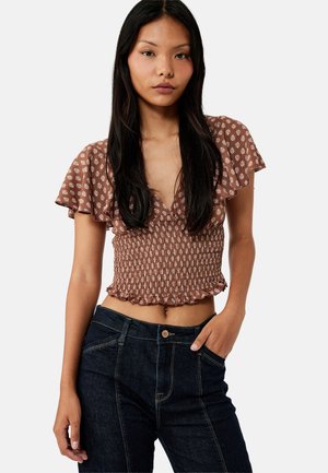 Frau mit langen schwarzen Haaren, die ein braunes gemustertes gerafftes Cropped-Top und dunkle High-Waist-Jeans trägt, steht mit einer Hand in der Tasche.