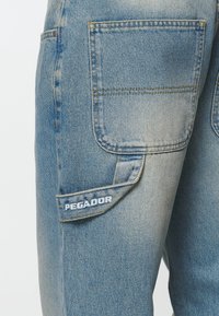 Ljusblå denimbyxor med en sydd fylld ficka med texten "PEGADOR." Tyget har en urtvättad textur med kontrasterande gula sömmar.