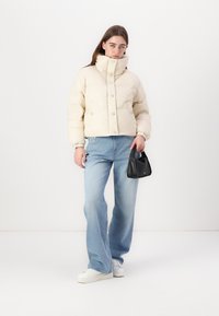 Doudoune crème avec col haut et fermetures à boutons, associée à un jean large bleu clair et un petit sac à main noir. Baskets blanches.