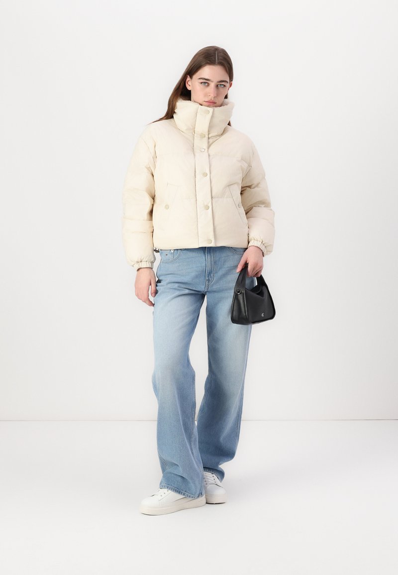 Cremefarbene Pufferjacke mit hoch geschlossenem Kragen und Knopfleiste, kombiniert mit hellblauen weit geschnittenen Jeans und einer kleinen schwarzen Handtasche. Weiße Sneaker.