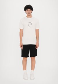 Hvit bomull t-skjorte med svart logo design, kombinert med sorte shorts og hvite joggesko. Modellen står rett mot en ensfarget bakgrunn.