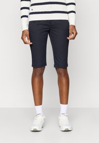 Calções azul-marinho com um corte ajustado, com bainha dobrada e bolsos laterais, combinados com tênis brancos e um suéter canelado com riscas azul-marinho.