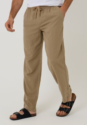 Mand iført beige snørede bukser og sorte båndsandal, stående med den ene hånd i lommen mod ensfarvet baggrund.