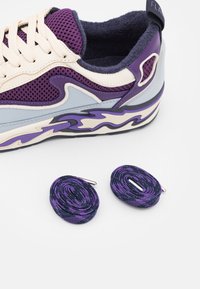 Baskets violettes et crème avec panneaux en mesh et semelle au motif ondulé, accompagnées de deux lacets roulés violets et noirs sur fond blanc.