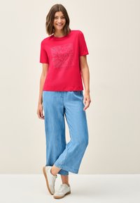 Rød bomulds T-shirt med grafisk print, parret med lyseblå bredbenede bukser. Fodtøj: hvide sneakers med en tan sål.