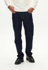 Vaqueros de mezclilla azul oscuro con corte recto, cinco bolsillos y costuras amarillas. Llevados con zapatos negros con cordones y un suéter blanco.