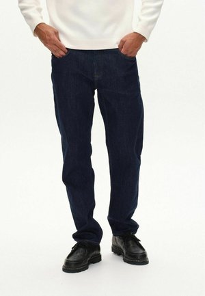 Vaqueros de mezclilla azul oscuro con corte recto, cinco bolsillos y costuras amarillas. Llevados con zapatos negros con cordones y un suéter blanco.