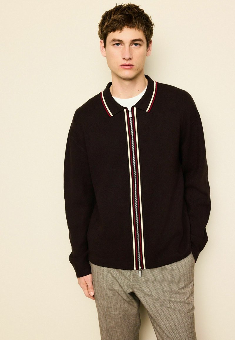 Cardigan zippé noir avec col et accents rayés rouges/blancs. Texture tricotée avec une coupe décontractée, associé à un pantalon à carreaux clair.