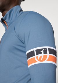 Zils sporta tops ar augstu kaklu, ar oranžu rāvējslēdzēja detaļu. Piedurkne izceļ logotipu un daudzkrāsu lentu ar melnām, baltām un oranžām sekcijām.