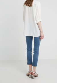 Blusa blanca de corte holgado con mangas enrolladas, combinada con jeans skinny de color azul oscuro y tacones de serpiente grises. Textura de tela suave.