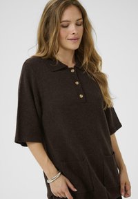 Brun, kortärmad, stickad polo med krage, fyra knappar i placket och två framfickor. Mjuk textur, avslappnad design.
