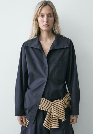 Femme aux longs cheveux blonds portant une chemise noire ample boutonnée et un pull rayé beige et noir noué autour de la taille.