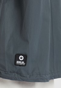 Szara wodoodporna kurtka wykonana z gładkiego materiału, z czarną prostokątną metką marki z białym logo przyszytym na dolnym końcu.