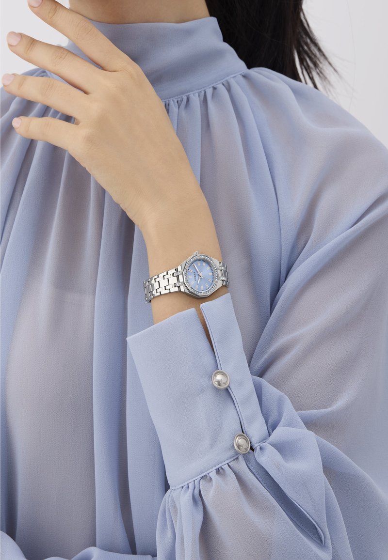 Hõbe kell, millel on helesinine kellapilt ja kristallaktsentidega bezel, kantud randmel, millel on helesinine pluus, millel on volangidega varrukad.