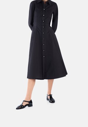 Black shirt dress with a fitted bodice, long sleeves, button front, and a flared skirt. Pair of black, strappy flat shoes with a block heel.