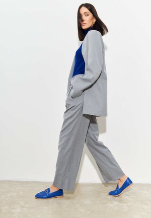Kvinde går, iført en lysegrå dragt, blå højhalset trøje og blå loafers, foran en ensartet hvid baggrund.