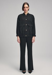 Zwarte jas met twee voorkepers, gouden knopen, elastische zoom, gecombineerd met zwarte flared broek en puntige zwarte hakken.