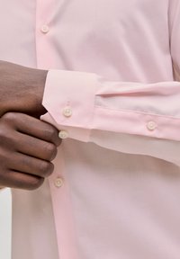 Ροζ πουκάμισο με μακριά μανίκια και λεία υφή, με λευκά κουμπιά και γωνιακά μανσέτα. Το ύφασμα φαίνεται μαλακό και ελαφρύ.